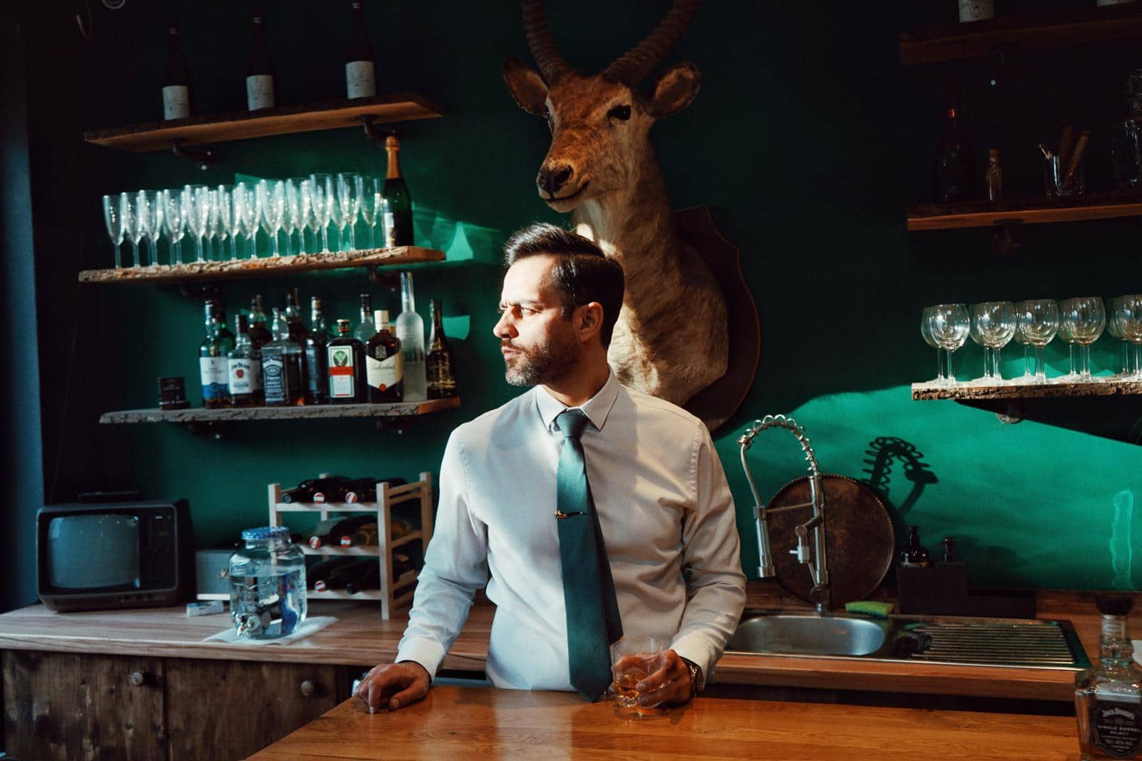 Shouka hinter der Bar seines Barbershops in Wien, vor dunkelgrüner Wand mit Wasserbock-Trophäe